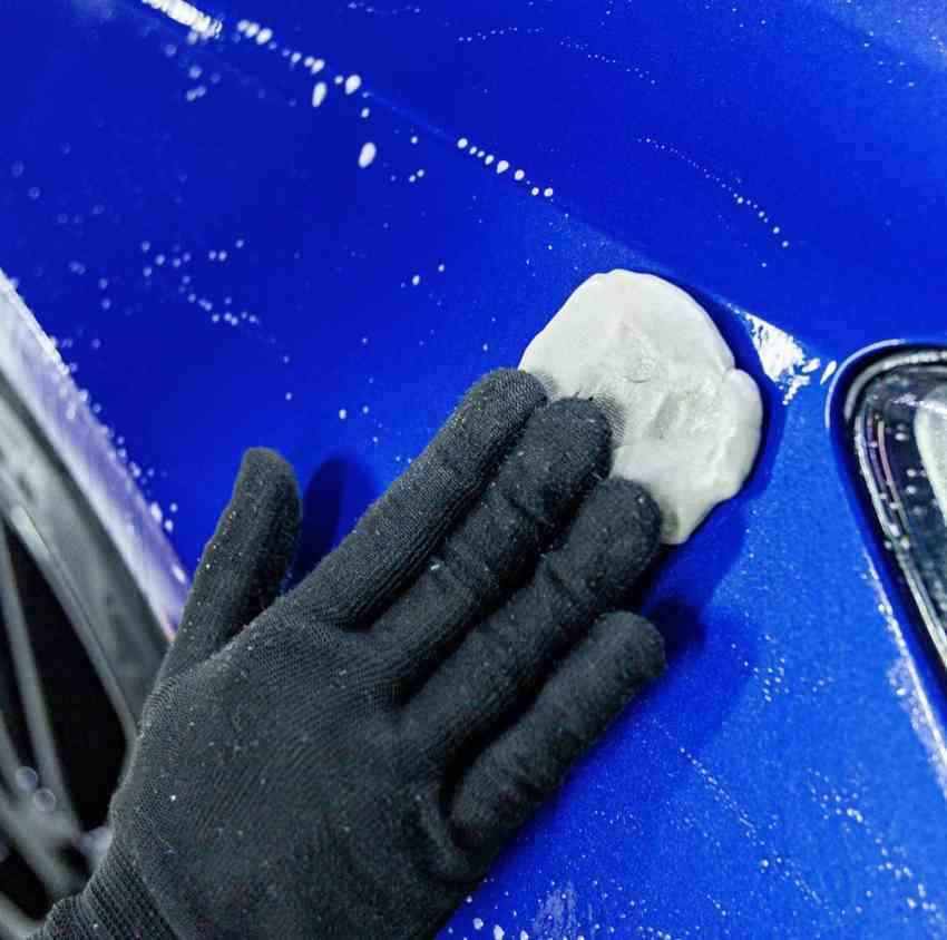 Hand with glove applying clay bar on blue car for paint correction treatment.