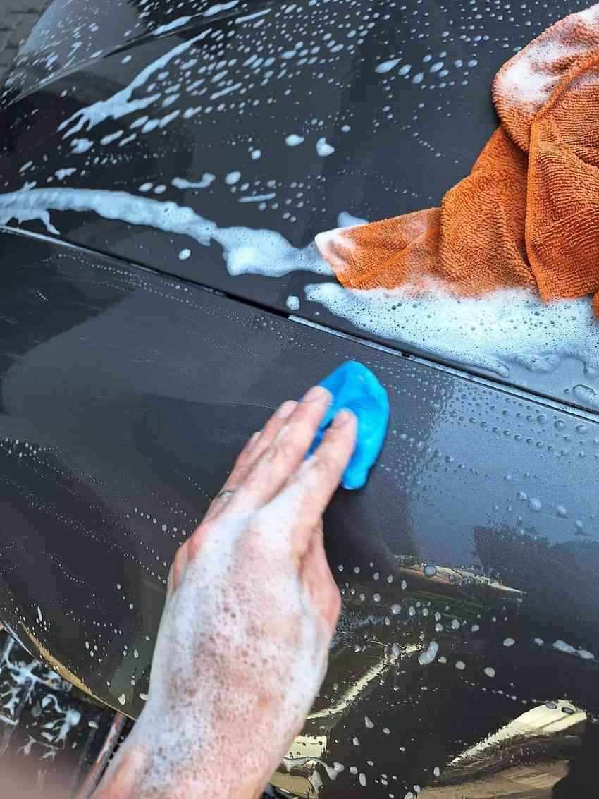 Hand applying clay bar treatment on car surface with soapy water during paint correction process.